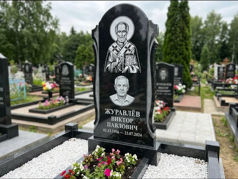 Памятник с гравировкой иконы: стела с образом в полный рост на обороте — гравировка образа Матроны Московской, низкие кусты спиреи в цвету, работа «Лебедева» в Пензе